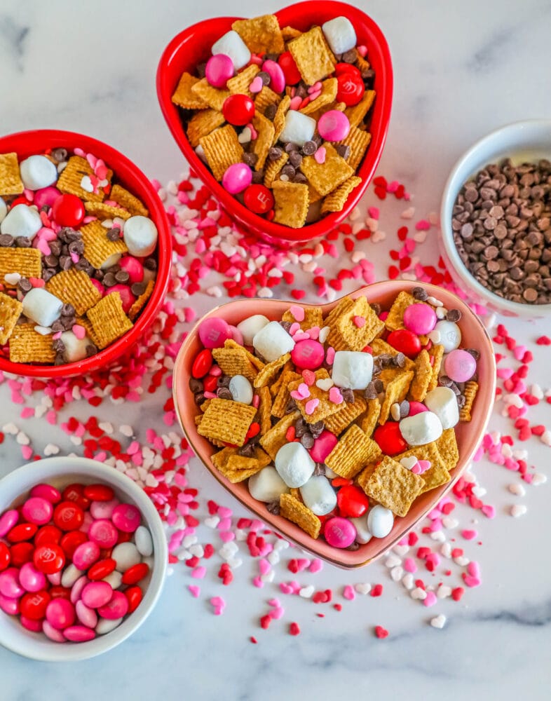 cereal, marshmallows, pink M&Ms, and chocolate chips in a bowl. Bowls of chocolate chips, sprinkles, and m&ms in the background