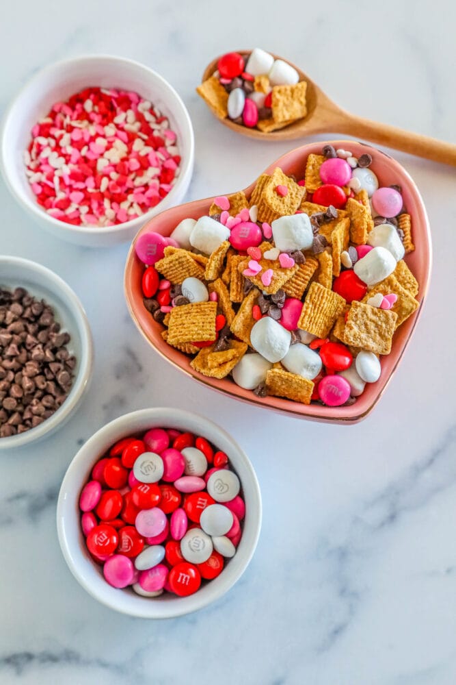 cereal, marshmallows, pink M&Ms, and chocolate chips in a bowl. Bowls of chocolate chips, sprinkles, and m&ms in the background