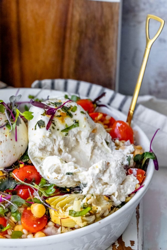 burrata barley salad in a white bowl with microgreens, artichoke hearts, and tomatoes