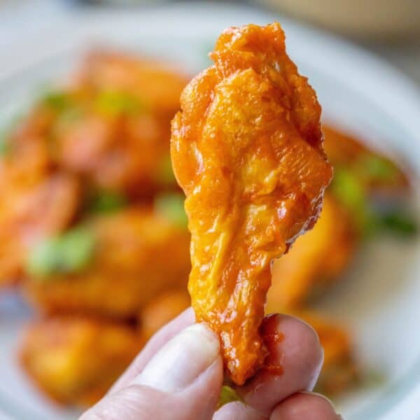 A hand holding a single baked buffalo wing with a blurred background of more crunchy baked chicken wings on a plate.