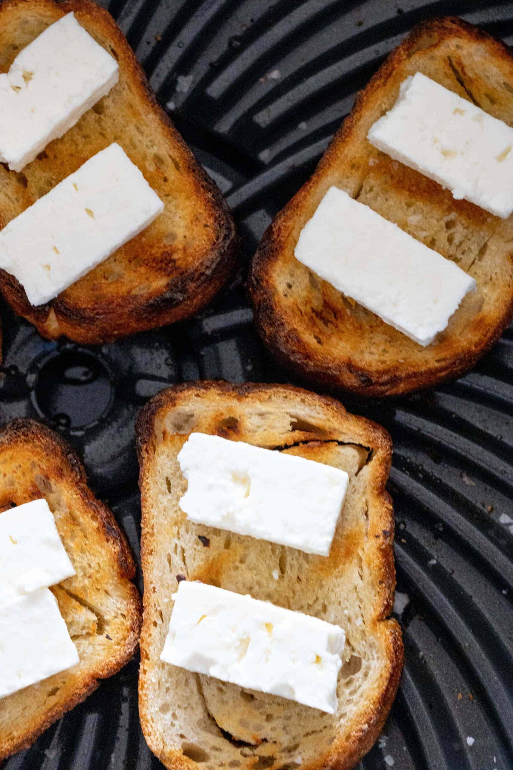 Four slices of toasted bread topped with rectangular pieces of white cheese, arranged on a dark ridged surface—perfect for trying a warm goat cheese salad or recreating a classic goat cheese salad recipe at home.
