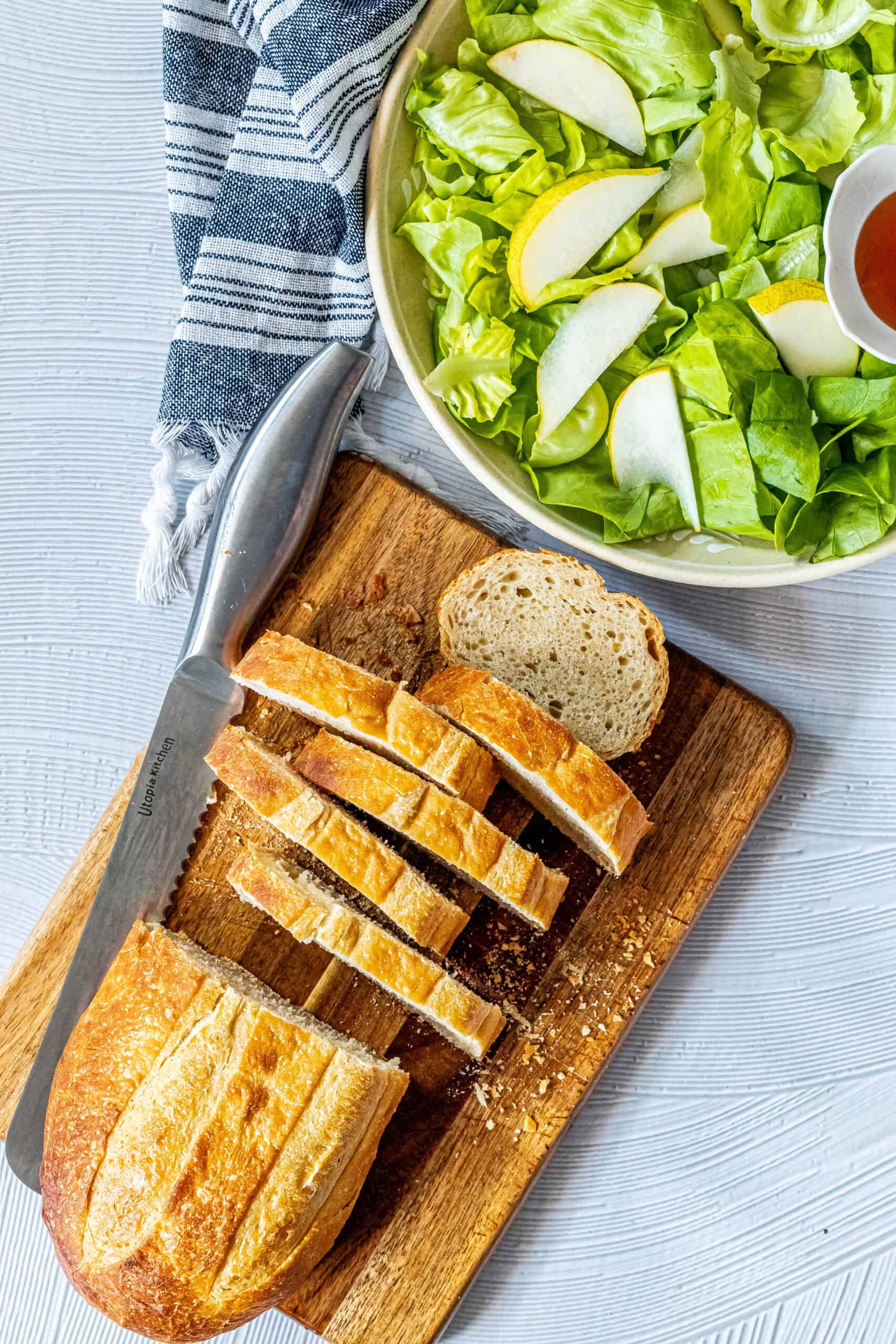 Sliced bread on a wooden cutting board with a knife beside a bowl of green salad topped with apple slices, inspired by a classic warm goat cheese salad, next to a striped cloth napkin.
