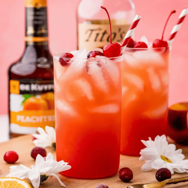 Two glasses of a vibrant Sex on the Beach Cocktail garnished with cherries and cranberries, striped straws, peach liqueur and vodka bottles, flowers, and fresh fruitโall you need for easy Sex on the Beach drinks on a wooden surface.