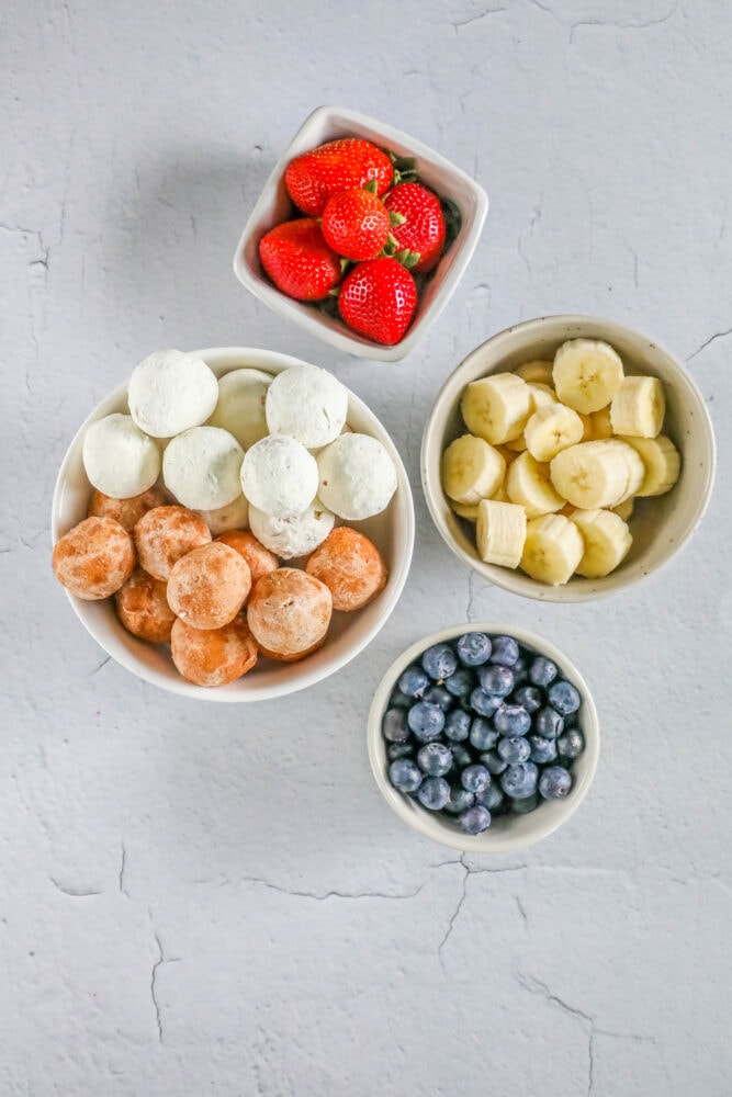 picture of a bowl of strawberries, a bowl of blueberries, a bowl of sliced bananas, and a bowl of donut holes on a table