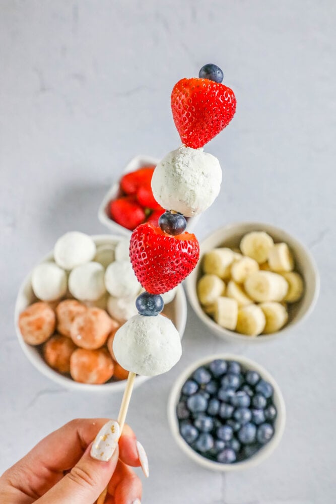 picture of a hand holding a kabob with a donut hole, blueberries, and strawberries on it