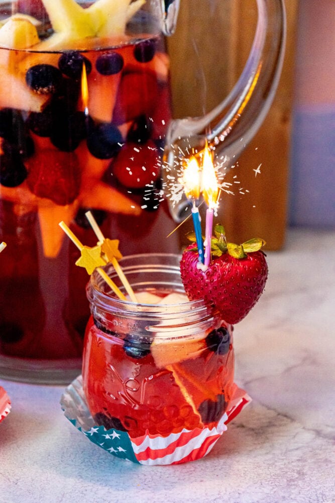 fruit punch with red white and blue fruit in it and a sparkling candle sticking out of a strawberry on the rim of the glass