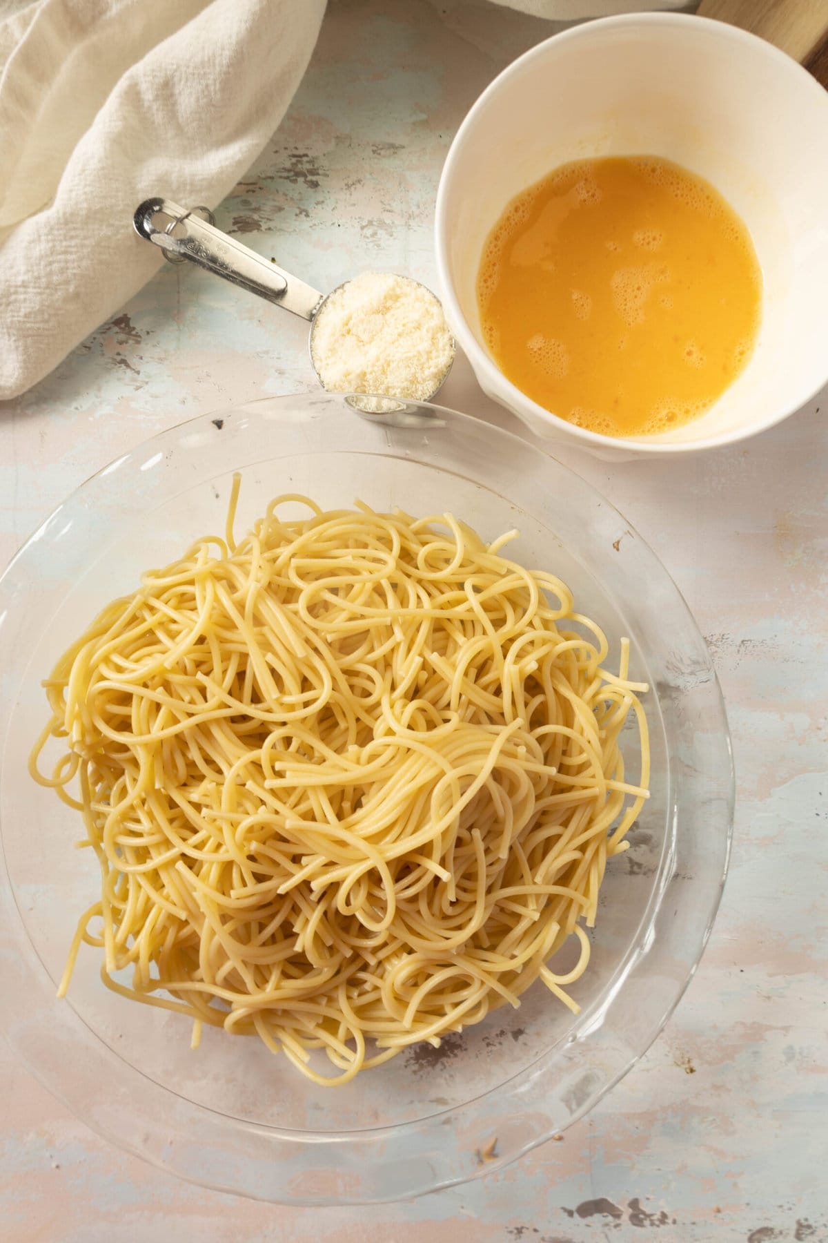 picture of boiled spaghetti in a pie plate with parmesan cheese and eggs on a table