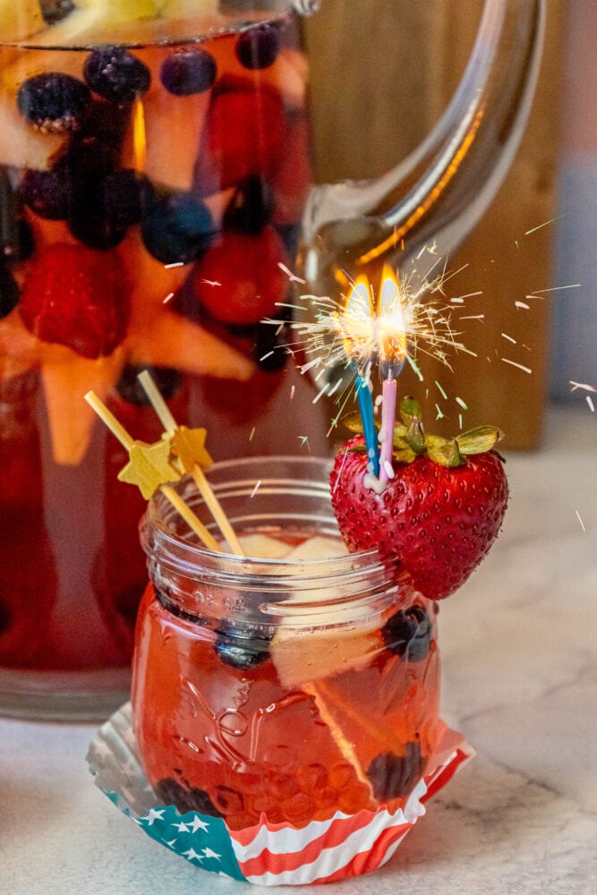 fruit punch with red white and blue fruit in it and a sparkling candle sticking out of a strawberry on the rim of the glass