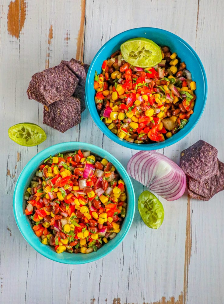 picture of salsa with beans, pepper, onion, corn, and herbs in two blue bowls on a table with chips