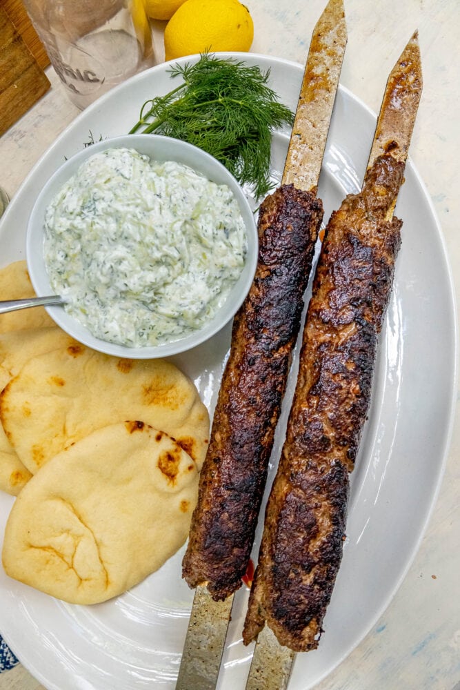 picture of two Greek gyros on skewers on a plate with tzatziki and pita bread on it