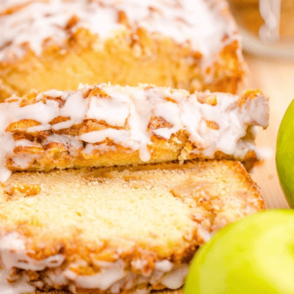 A close-up of a sliced loaf of sweet iced apple bread with icing on top, next to whole green apples.