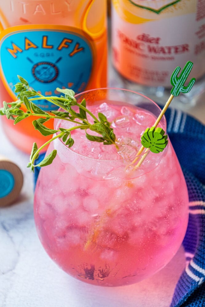 picture of pink gin and tonic in a large glass on a table with ice in it in front of a bottle of malfy con arancia gin