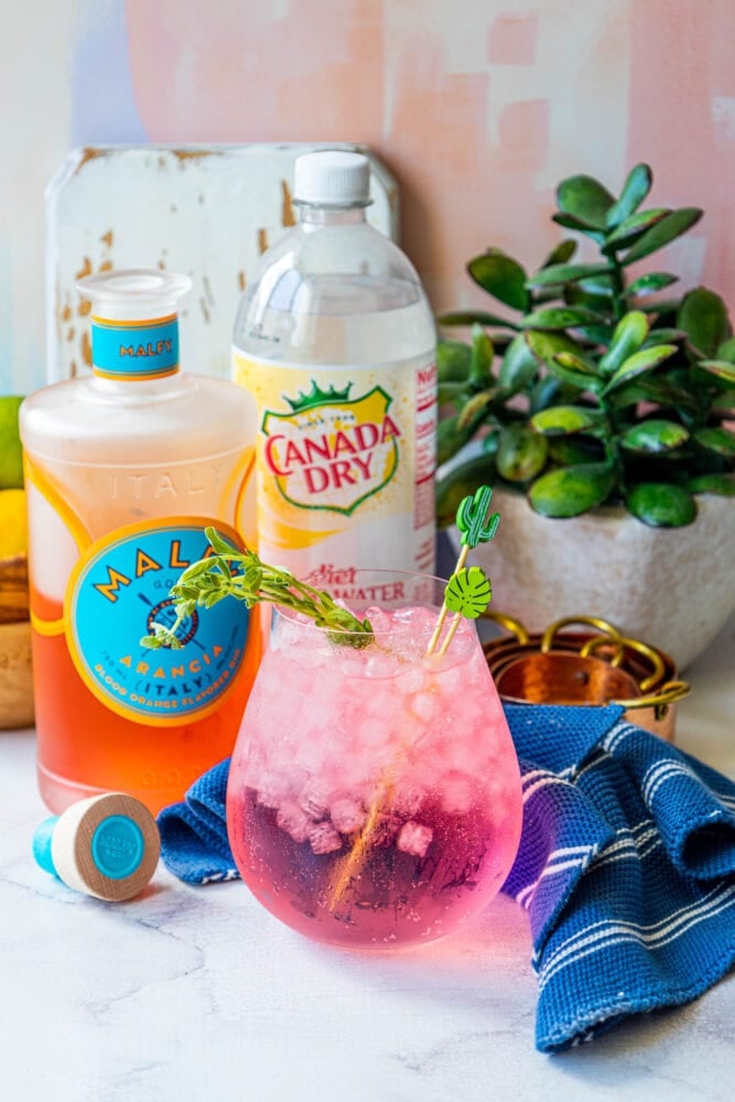 picture of pink gin and tonic in a large glass on a table with ice in it in front of a bottle of malfy con arancia gin