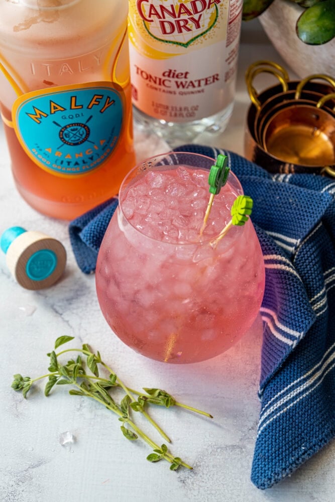 picture of pink gin and tonic in a large glass on a table with ice in it in front of a bottle of malfy con arancia gin