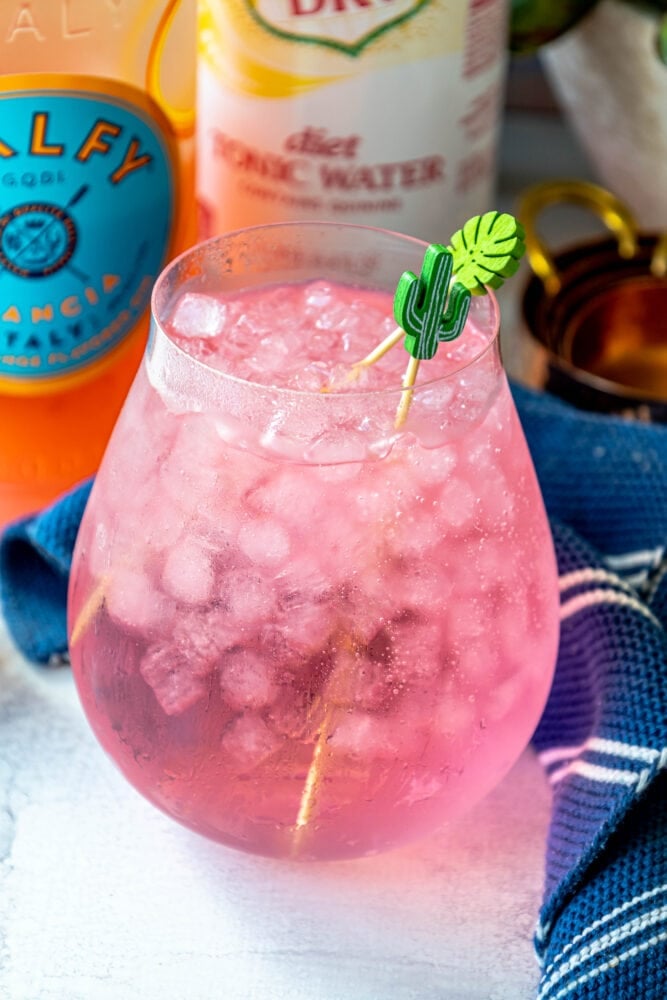 picture of pink gin and tonic in a large glass on a table with ice in it in front of a bottle of malfy con arancia gin