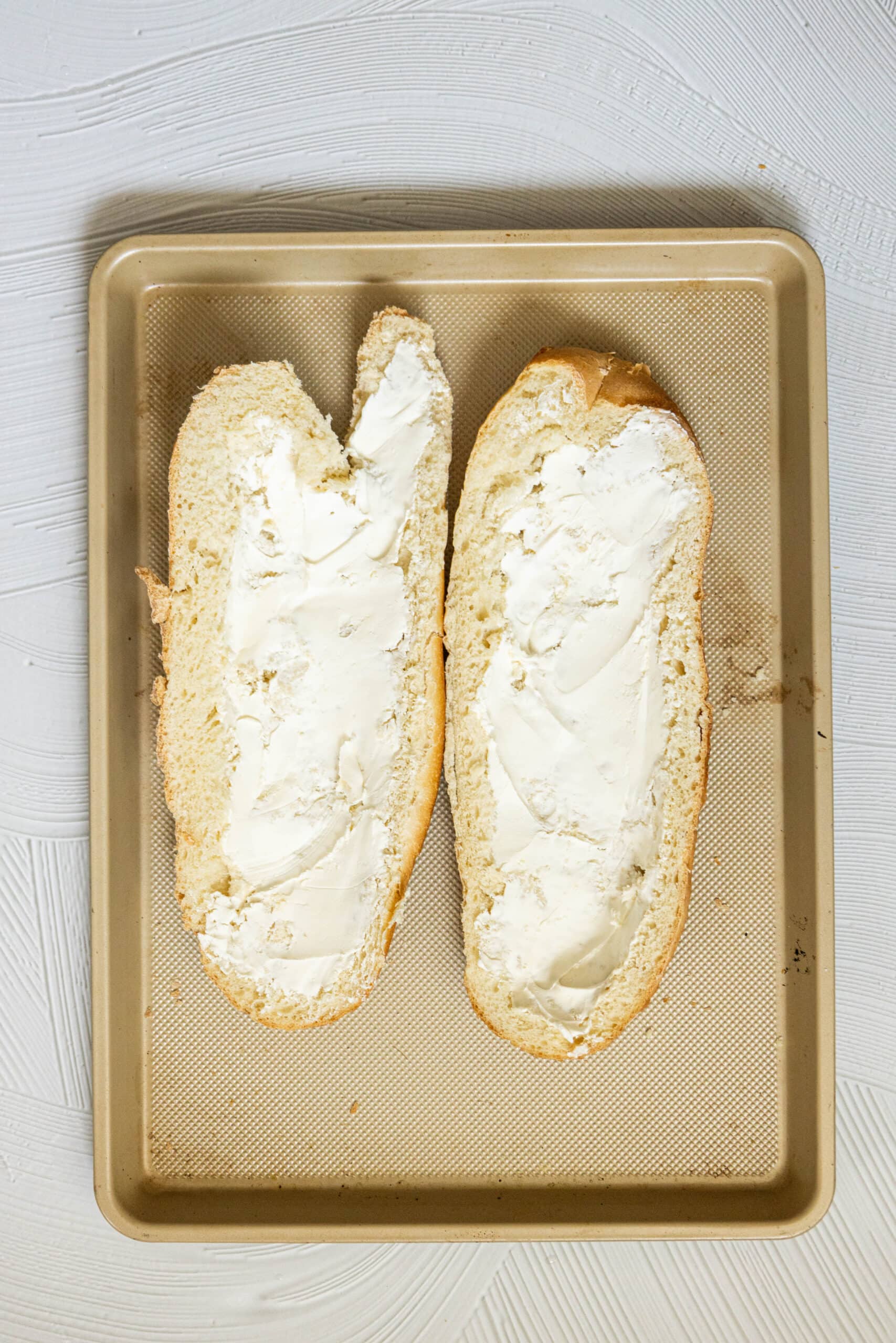 picture of cream cheese spread over two halves of french bread on a baking sheet