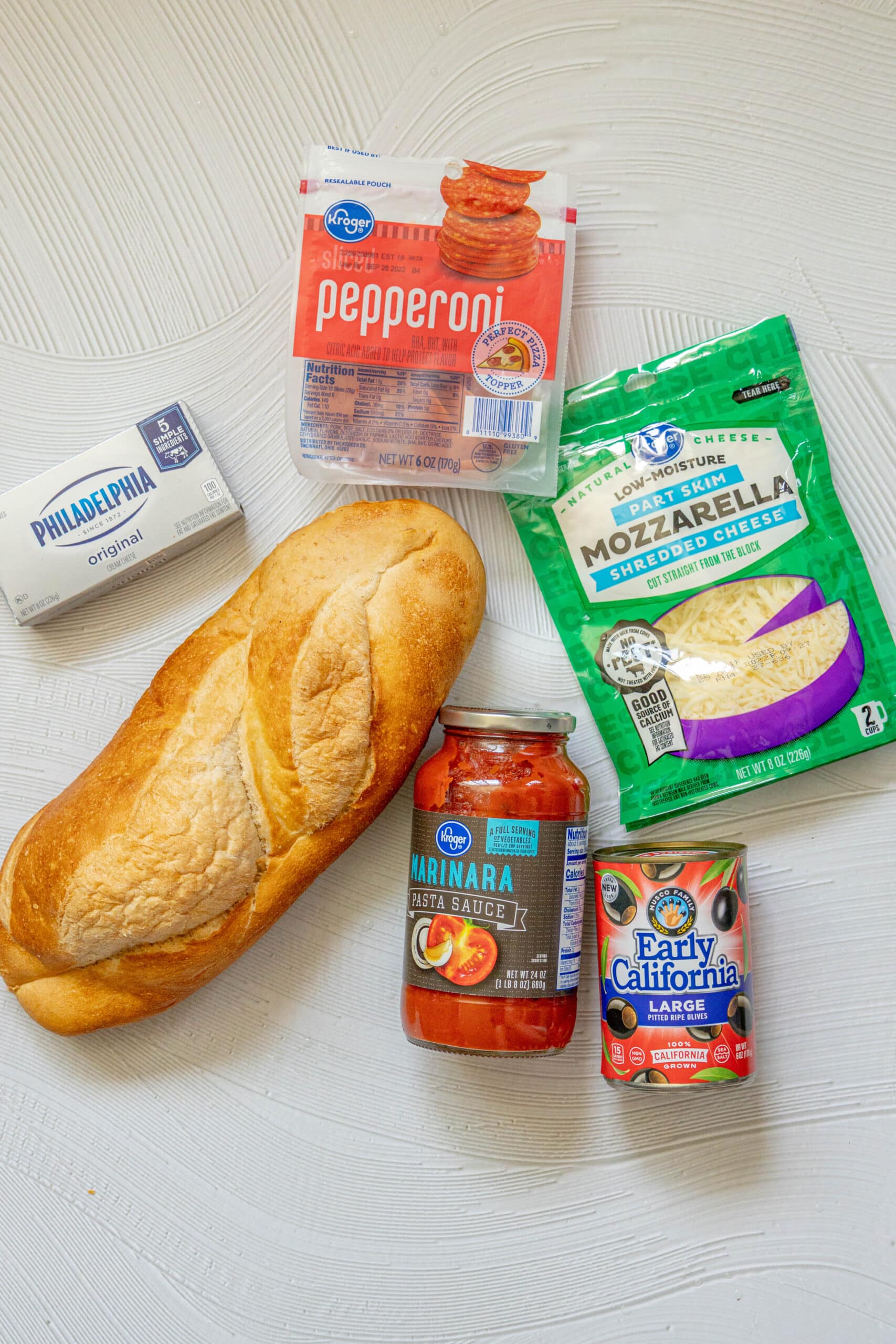 picture of pepperoni, cream cheese, french bread, marinara sauce, jarred olives, shredded mozzarella on a table