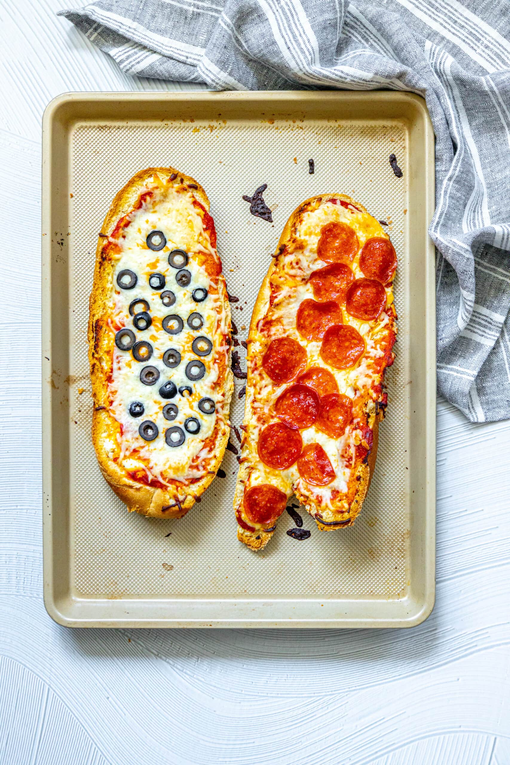 picture of french bread pizza cut in half on a baking sheet baked with cheese, pepperoni, and olives on top