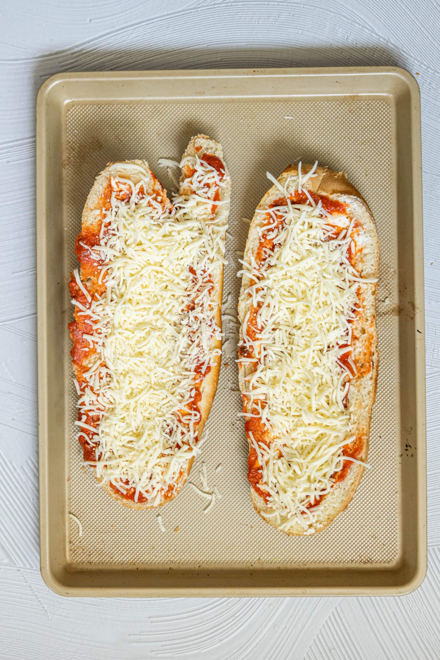 picture of cream cheese, sauce, and shredded mozzarella spread over two halves of french bread on a baking sheet
