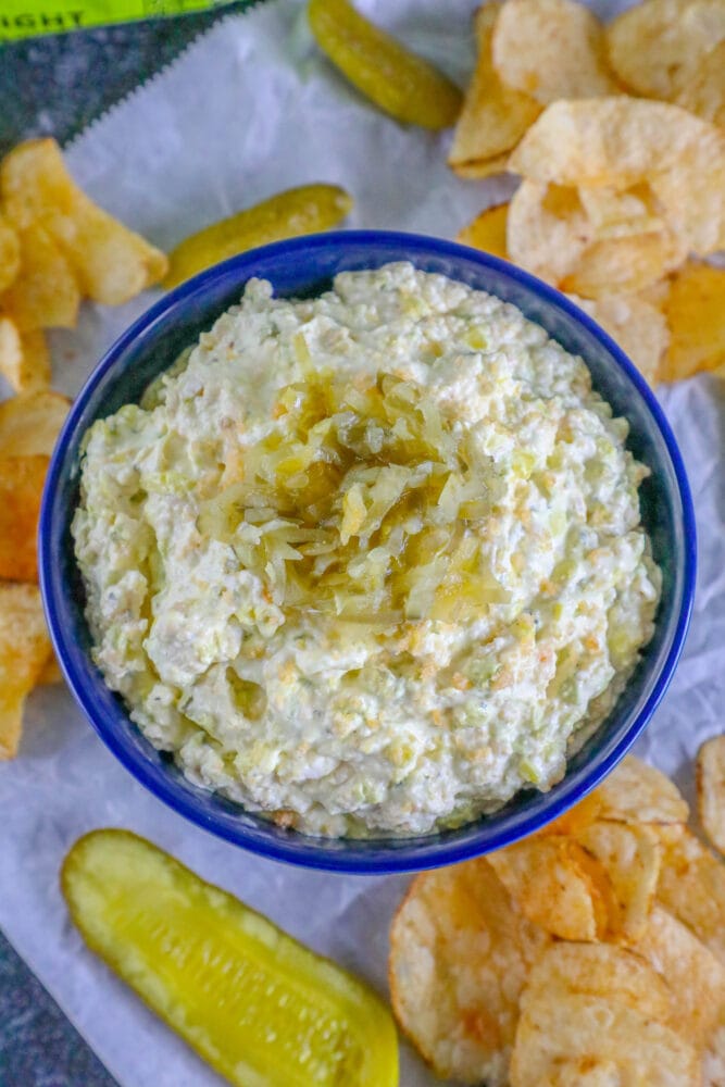 picture of pickle dip in a blue bowl around chips and a jar of pickles