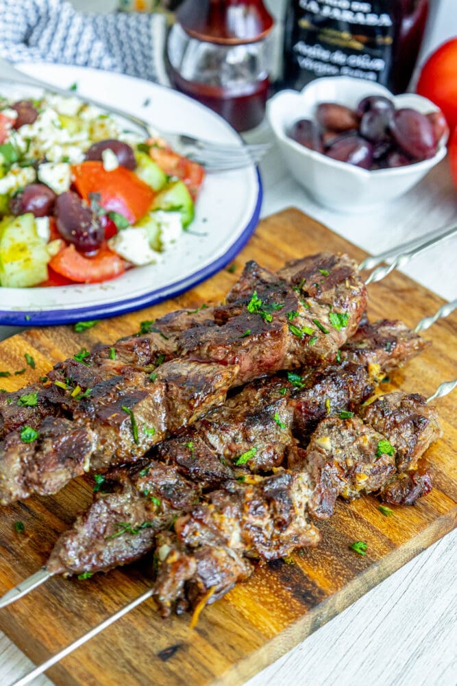 picture of lamb souvlaki on a skewer on a wood cutting board with chopped cilantro on top in front of a greek salad