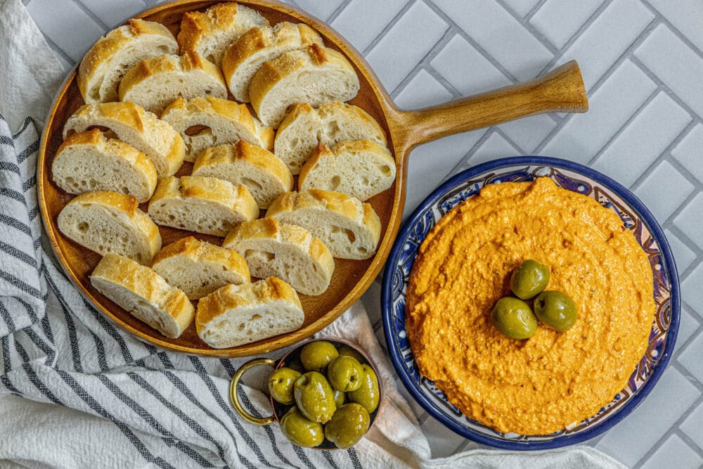 picture of whipped feta cheese dip with three olives on top in a blue bowl next to a bowl of bread and olives on a table