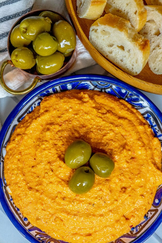 picture of whipped feta cheese dip with three olives on top in a blue bowl next to a bowl of bread and olives on a table