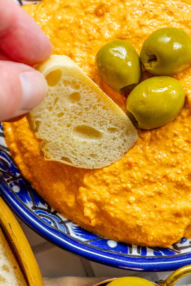 picture of hand dipping bread into whipped red pepper and feta cheese dip with olives on top in a blue bowl 