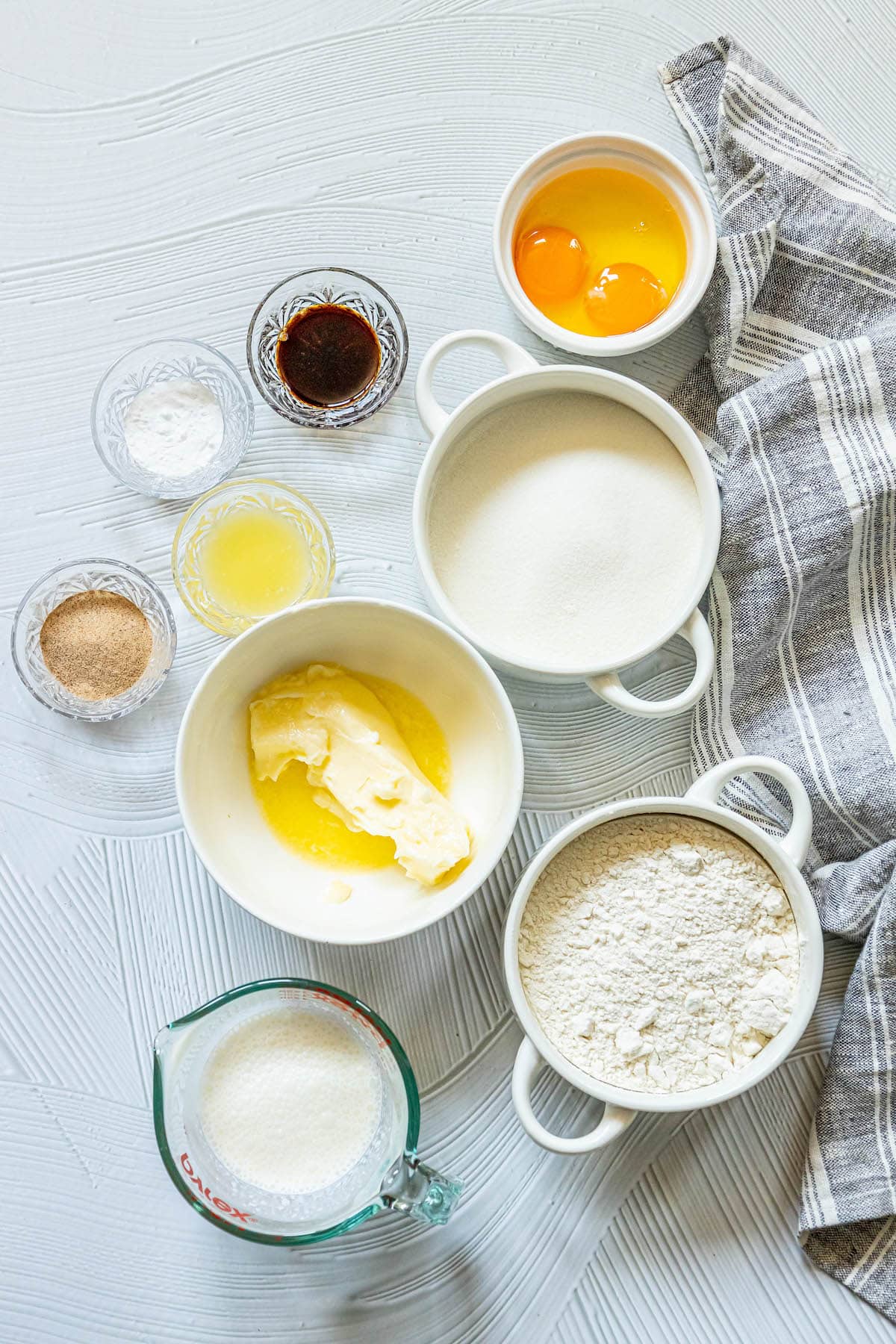 picture of vanilla, flour, sugar, eggs, butter, flour, sugar, salt, and milk in bowls on a table
