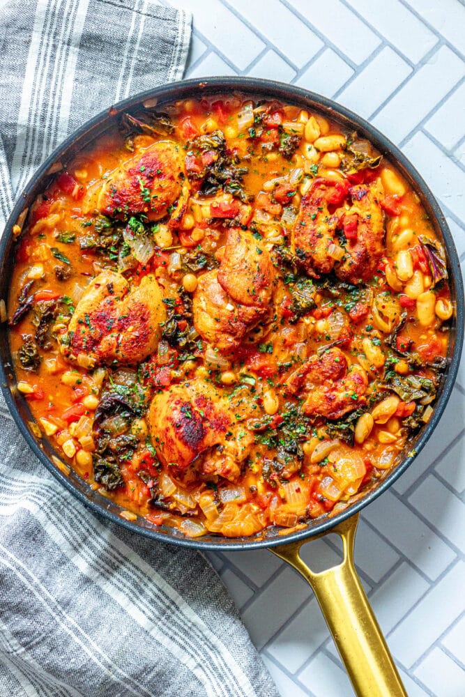 picture of six chicken thighs in tomato stew with white beans, kale, herbs, and onions in a pan on a table