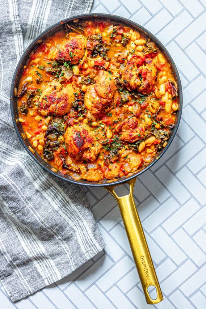 picture of chicken thighs in tomato stew with white beans, kale, herbs, and onions in a pan on a table