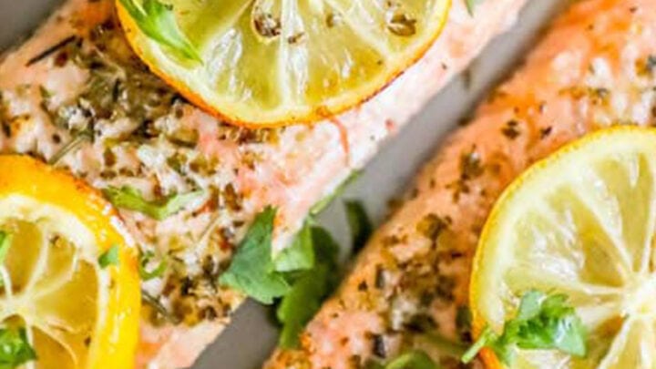picture of salmon filets with lemon slices, garlic and herbs on top on a white plate