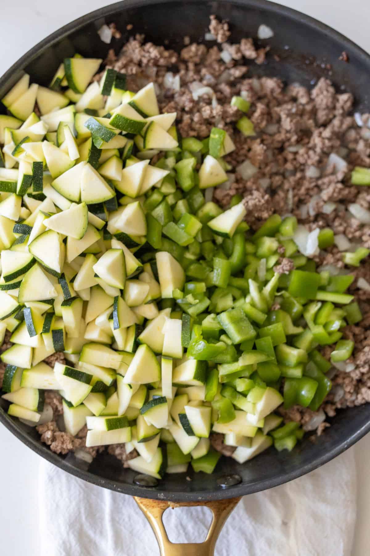A skillet filled with cooked ground beef and onions on one side, and raw chopped zucchini and green bell peppers on the other, ready to become a Cheesy Beef Taco Skillet.
