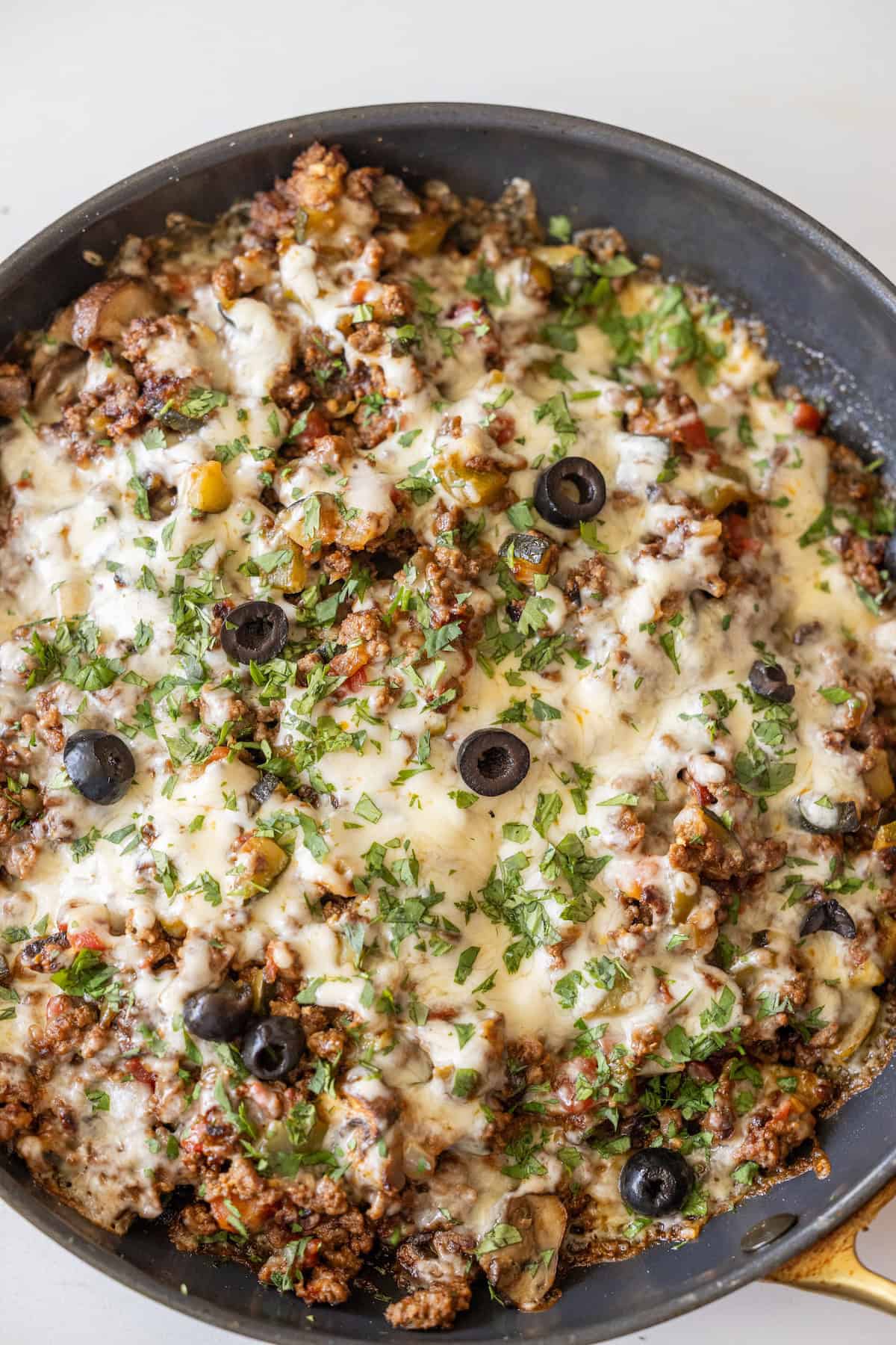 A Cheesy Taco Skillet filled with cooked ground meat, melted cheese, chopped herbs, black olive slices, and diced vegetables.