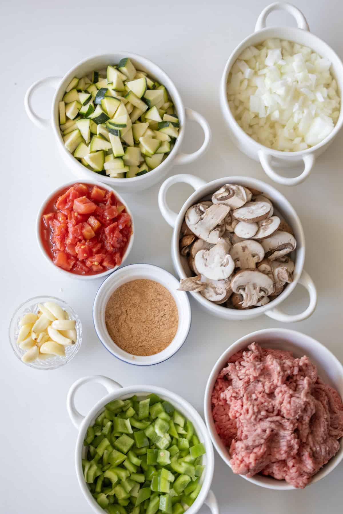 Several bowls filled with chopped zucchini, onions, tomatoes, mushrooms, green bell pepper, minced garlic, breadcrumbs, and ground meat are arranged on a white surface—perfect for preparing the Best Taco Skillet.