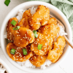 A bowl of chicken breast sesame chicken served over white rice, garnished with green onions and sesame seeds.