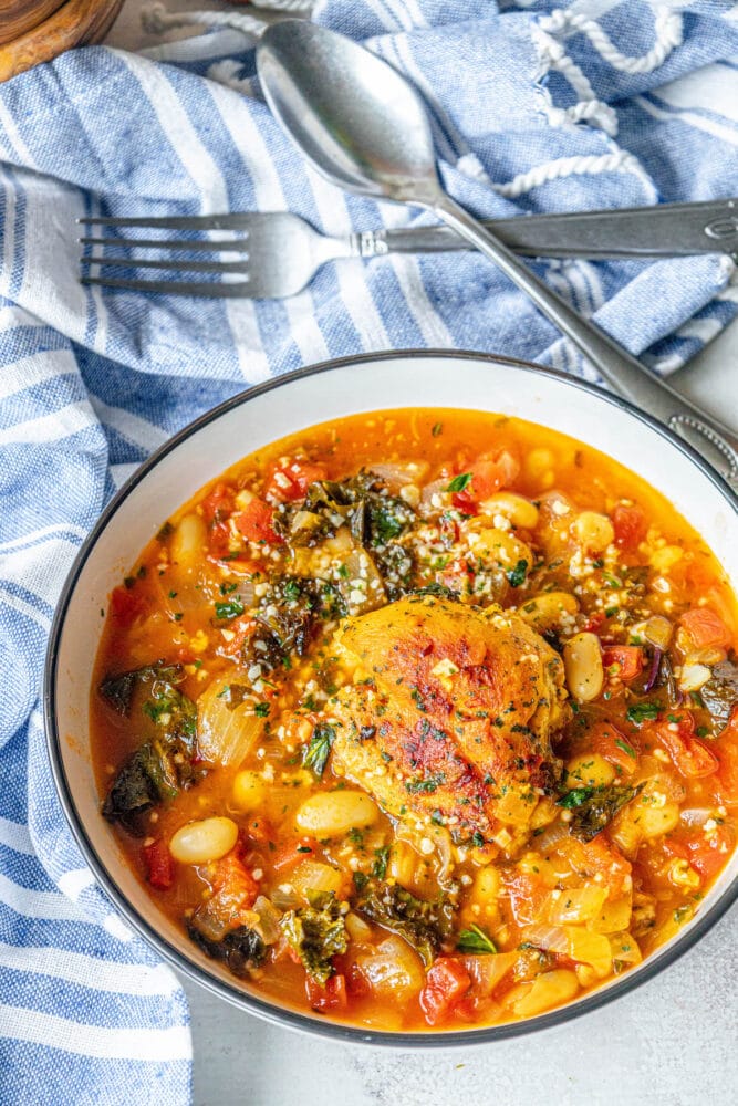 picture of chicken thigh in tomato stew with white beans, kale, herbs, and onions in a white bowl on a table with a towel and fork and spoon next to it 