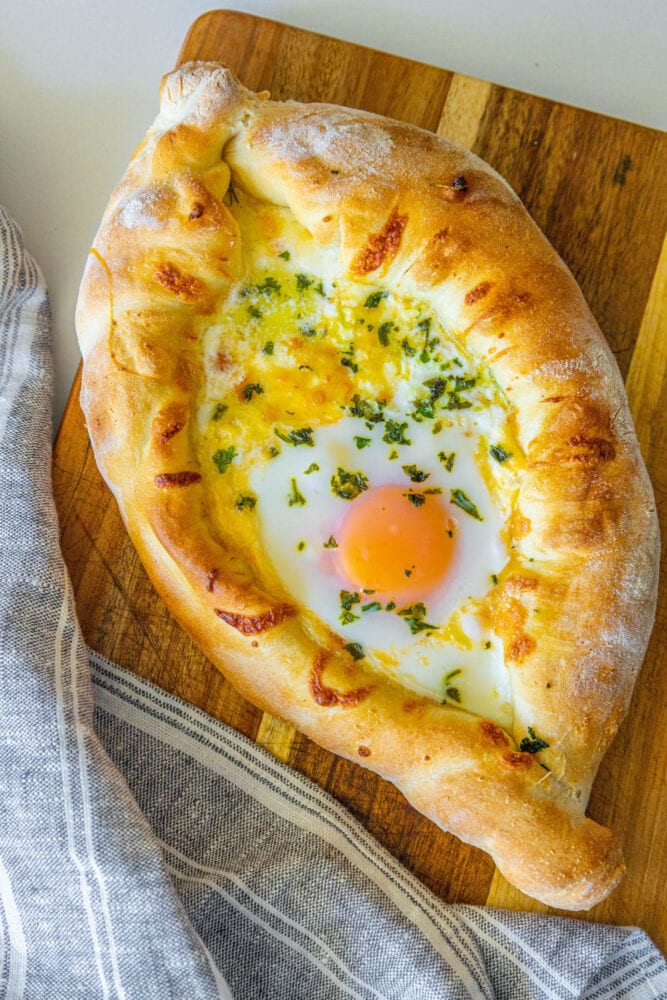 picture of baked khachapuri cheese bread on a wood cutting board with egg yolk and herbs on top 