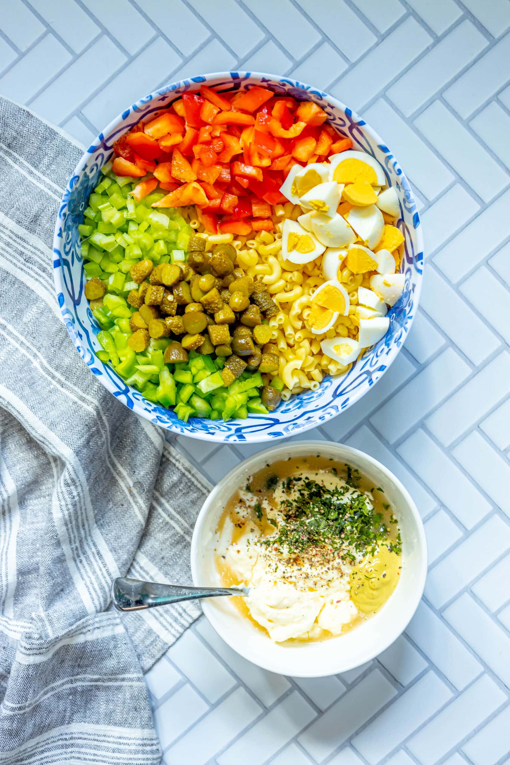 picture of cooked macaroni in a bowl with diced eggs, peppers, pickles, celery and peas on top next to a bowl of mayonnaise, mustard, vinegar, herbs, and spices on a table