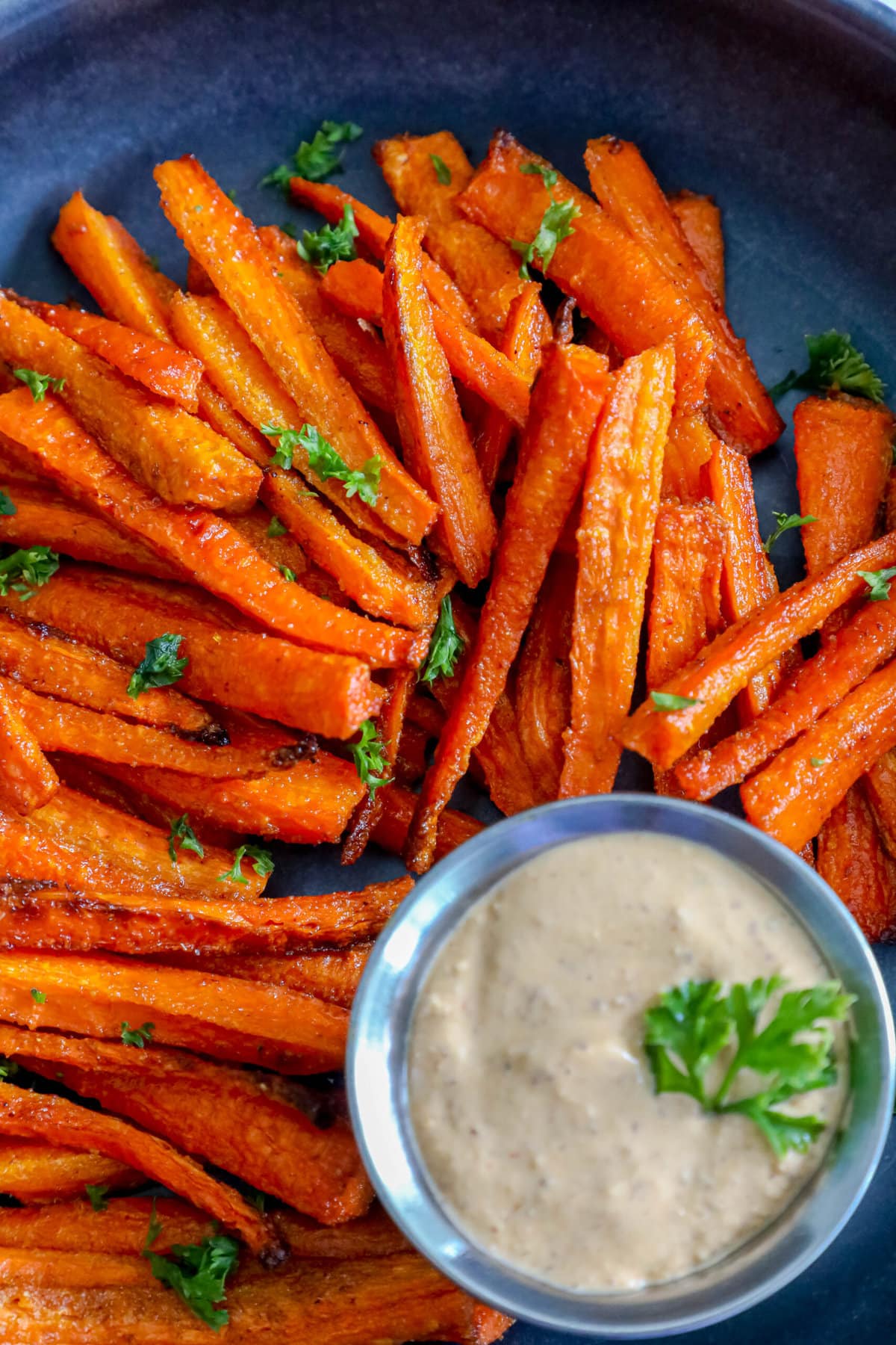 picture of carrot fries with parsley chopped on top on a black plate with a cup of sauce on the side