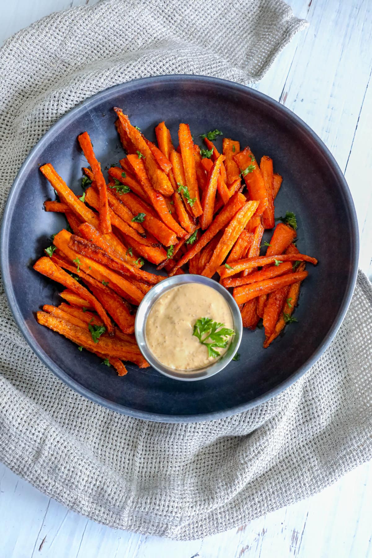 picture of carrot fries with parsley chopped on top on a black plate with a cup of sauce on the side