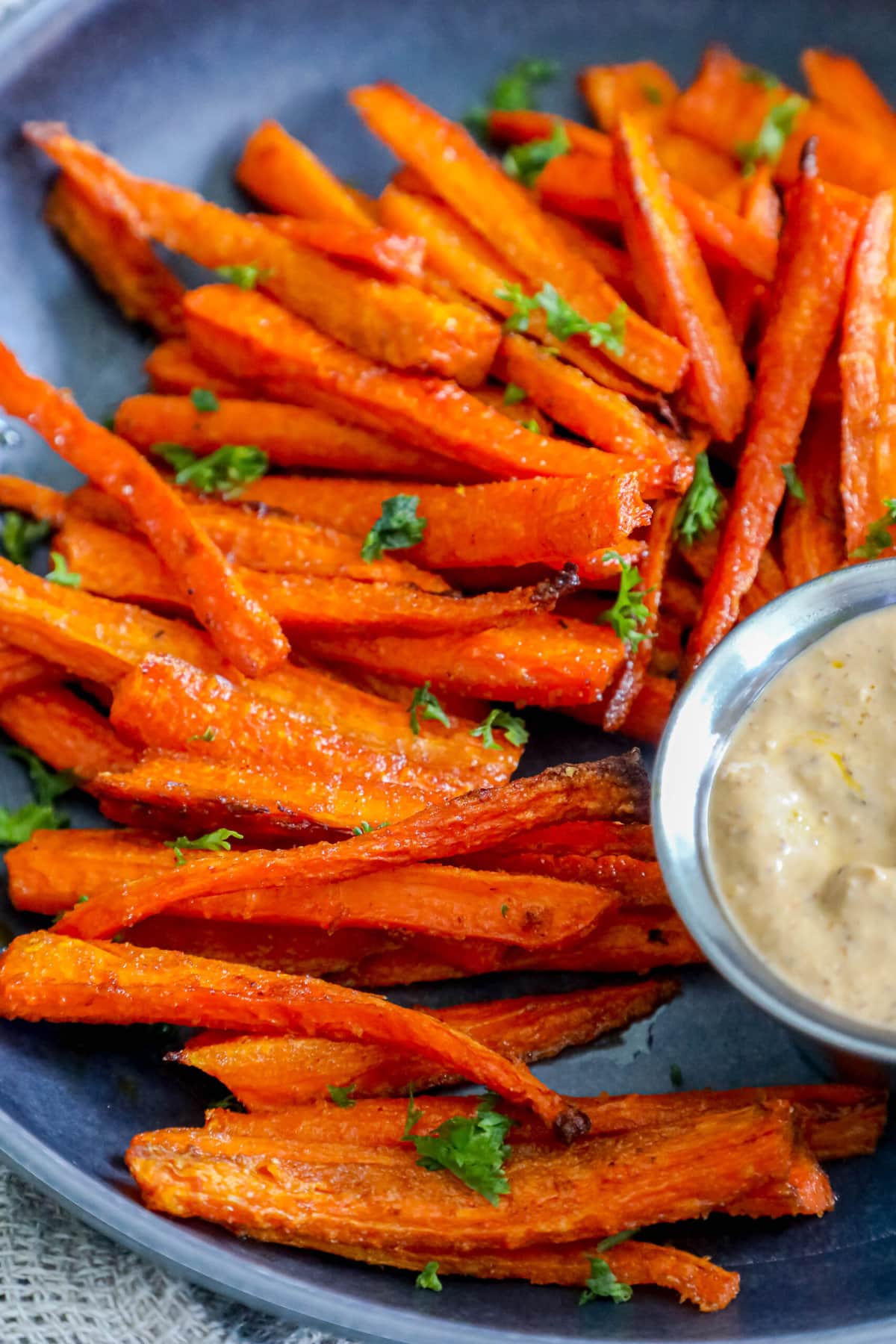 picture of carrot fries with parsley chopped on top on a black plate with a cup of sauce on the side