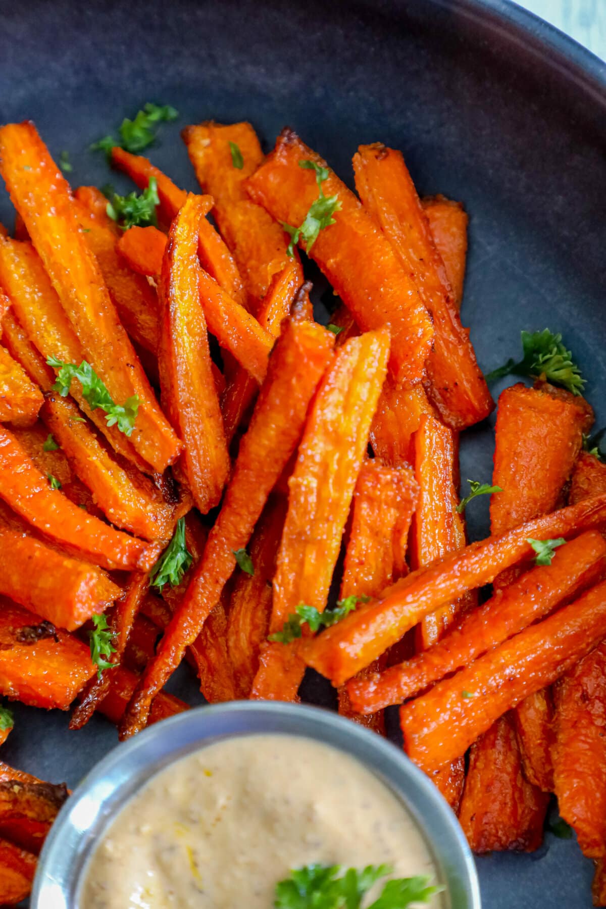 picture of carrot fries with parsley chopped on top on a black plate with a cup of sauce on the side