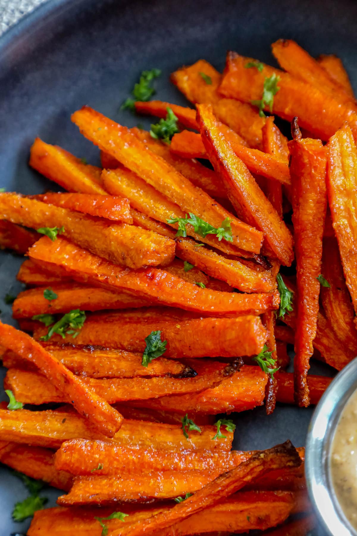 picture of carrot fries with parsley chopped on top on a black plate with a cup of sauce on the side