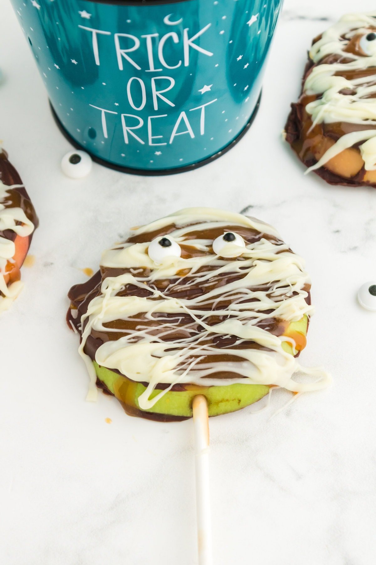 picture of caramel apples decorated as mummies with chocolate and candy eyes in a blue trick or treat bucket