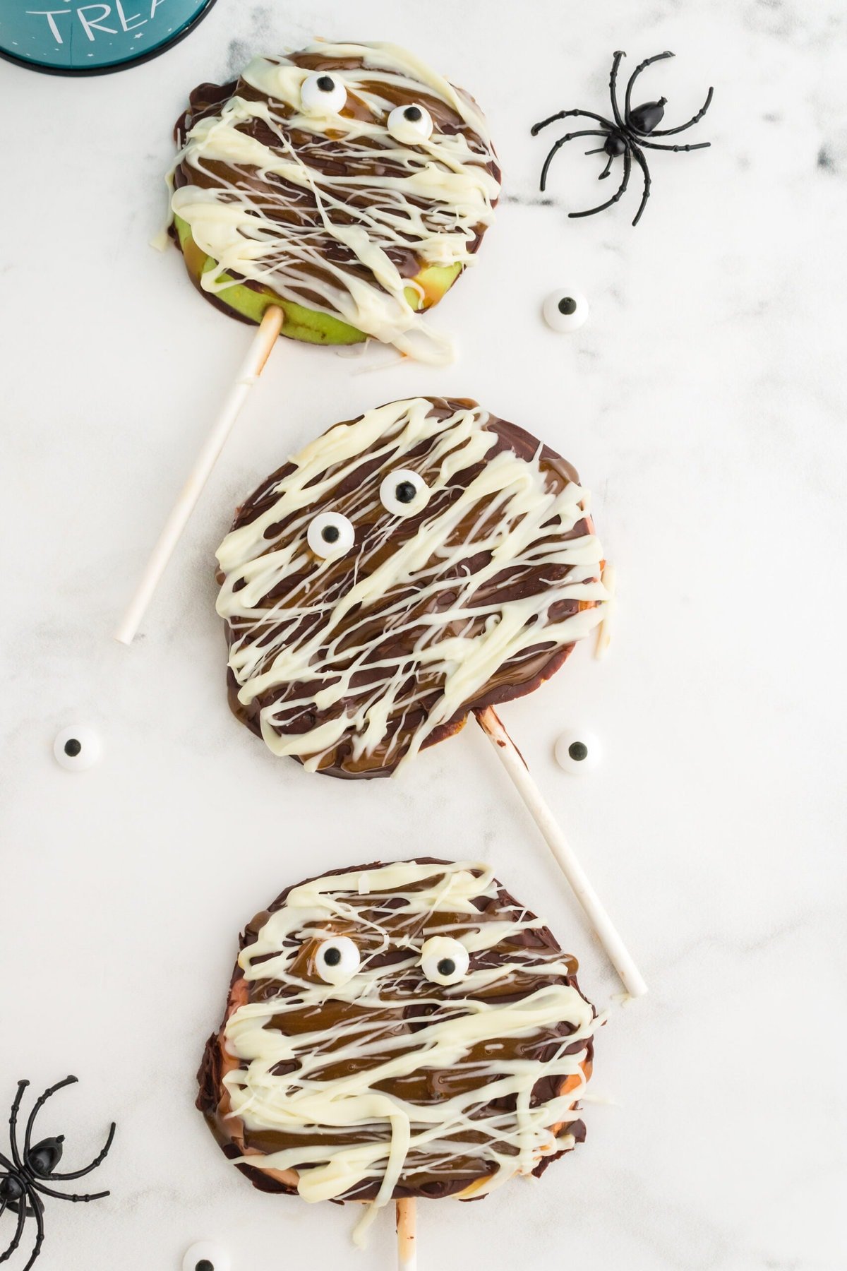 picture of caramel apples decorated as mummies with chocolate and candy eyes in a blue trick or treat bucket