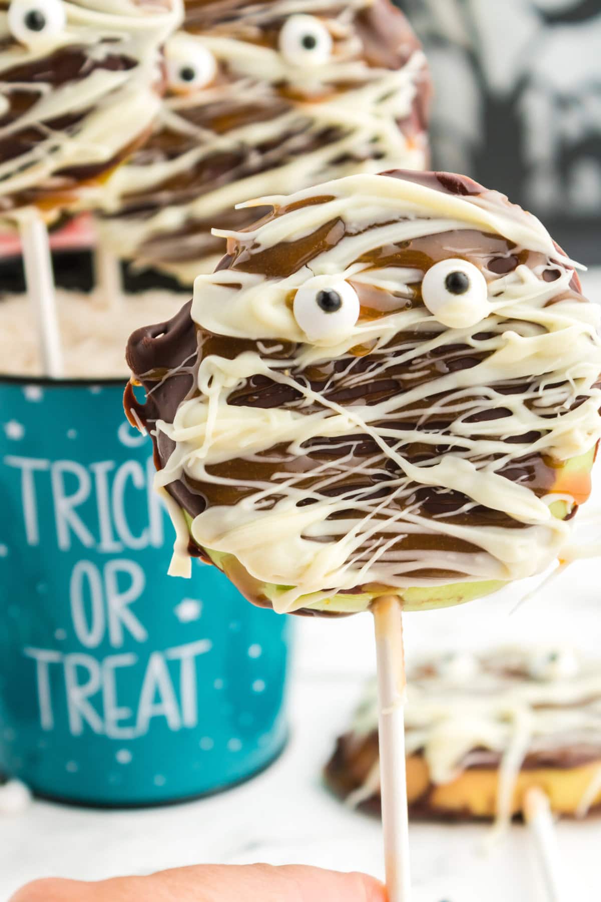 picture of caramel apples decorated as mummies with chocolate and candy eyes in a blue trick or treat bucket