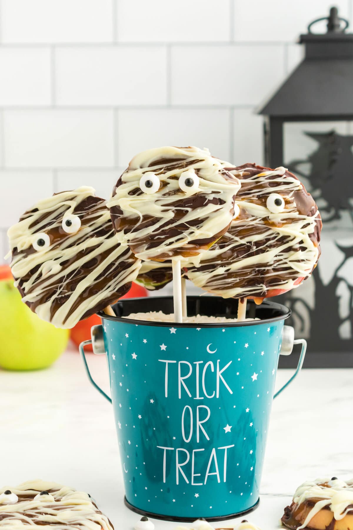 picture of caramel apples decorated as mummies with chocolate and candy eyes in a blue trick or treat bucket