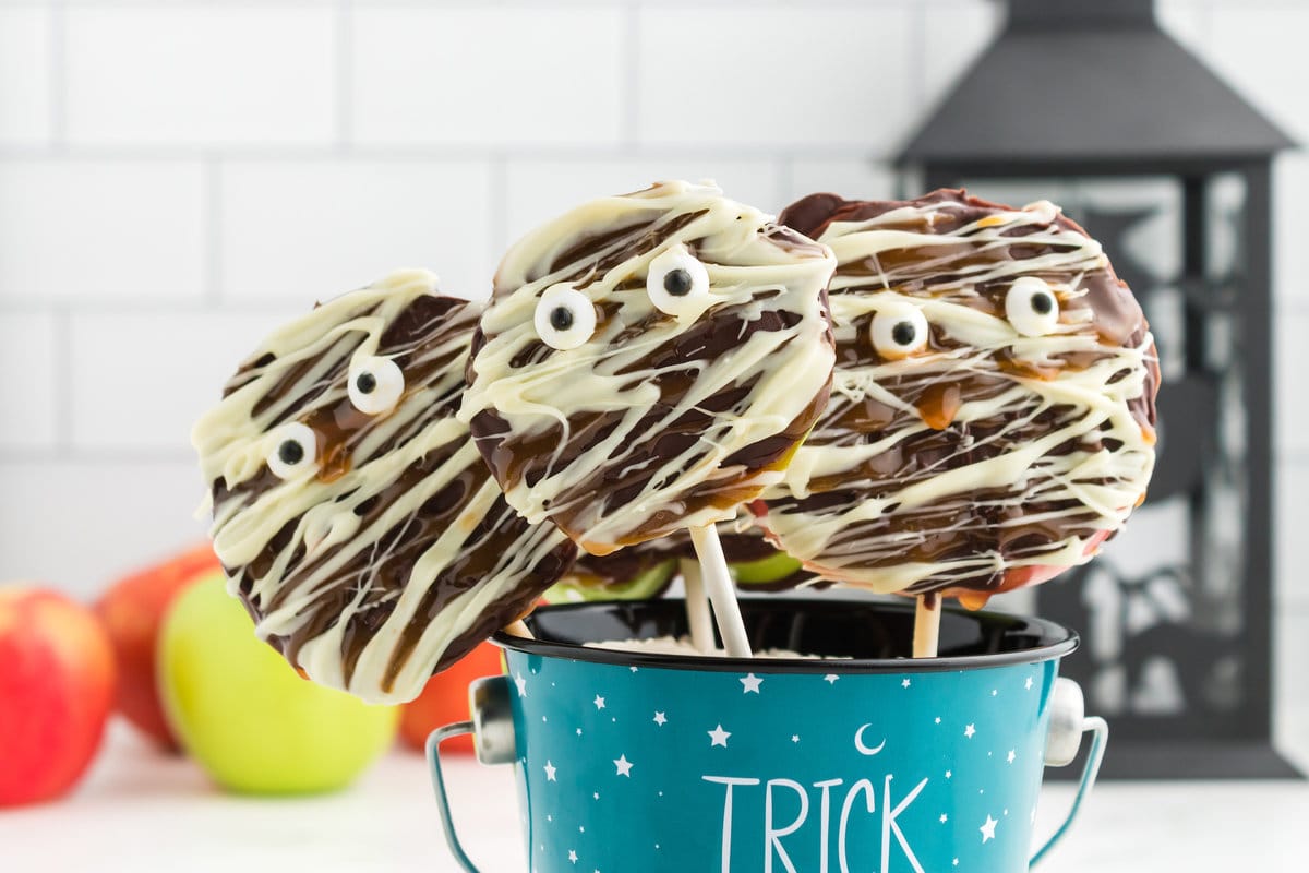 picture of caramel apples decorated as mummies with chocolate and candy eyes in a blue trick or treat bucket