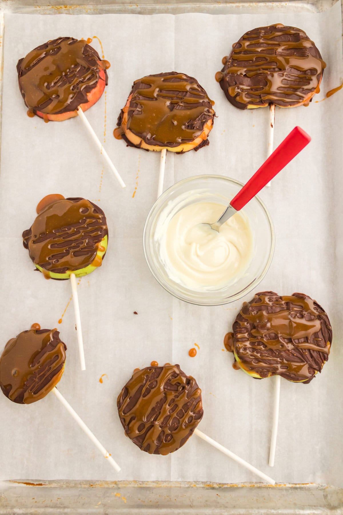 Caramel apple slices mummies dipped in chocolate on a baking sheet.